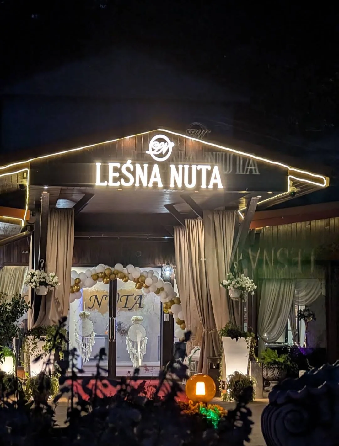 Oświetlone wejście do budynku Leśnej Nuty z kwiatami i dekoracjami nocą